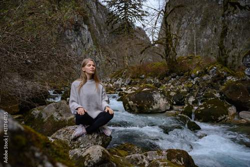 Girl sits on a large stone and meditates near a waterfall next to a mountain river.