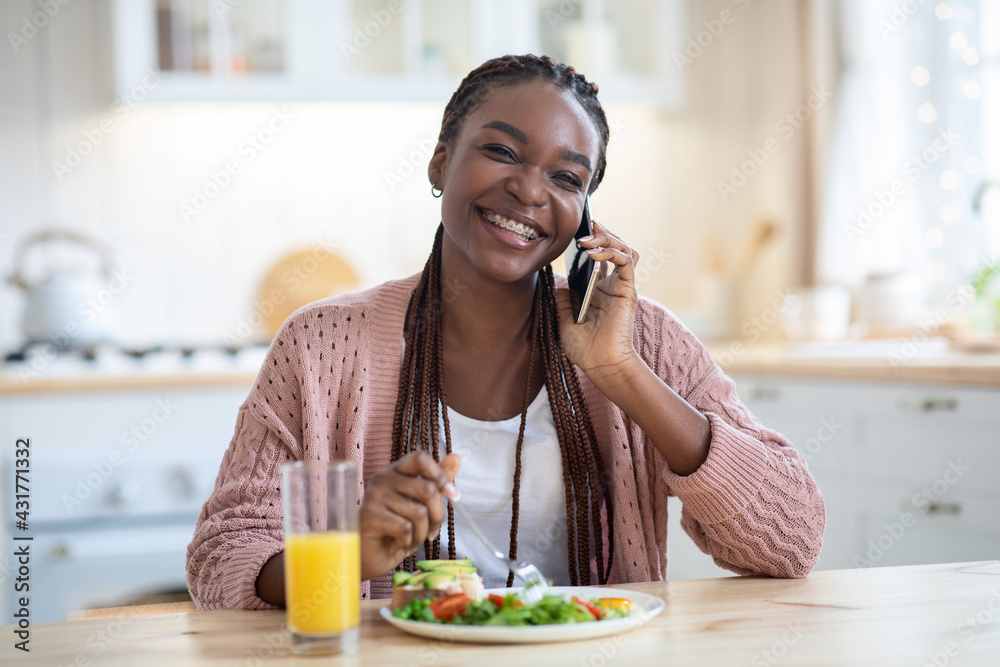 Happy Black Lady Talking On Mobile Phone While Eating Breakfast In Kitchen