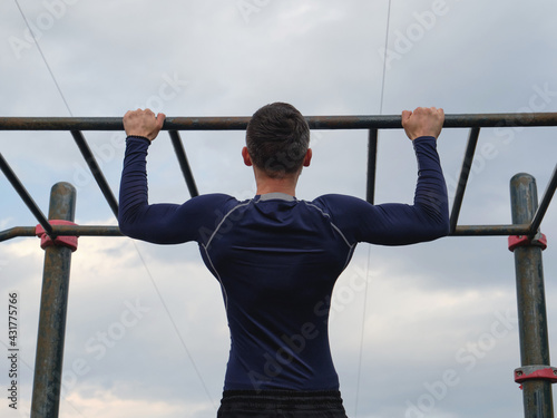 An athlete guy in a blue longsleeve pulls himself up on a horizontal bar. View from the back.