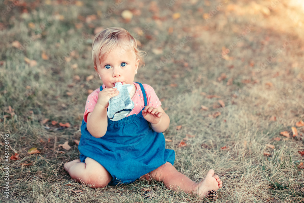 Toddler baby eating baby food organic vegetable fruit puree from pouch. Supplementary solid healthy food meal snack for babies kids. Girl eating outdoors in park on summer day. Toddler baby eating baby food organic vegetable fruit puree from pouch. Supplementary solid healthy food meal snack for babies kids. Girl eating outdoors in park on summer day.