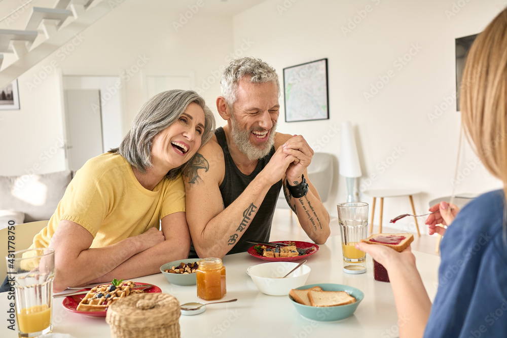 Happy mature family couple having breakfast with teen daughter sit at kitchen table. Cheerful older mid age parents having fun talking, eating toasts and waffles enjoying morning meal together.