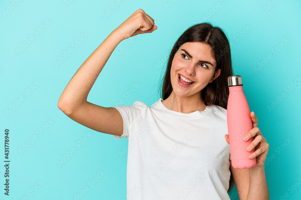 Young caucasian woman holding water bottle isolated on blue background raising fist after a victory, winner concept. Young caucasian woman holding water bottle isolated on blue background raising fist after a victory, winner concept.