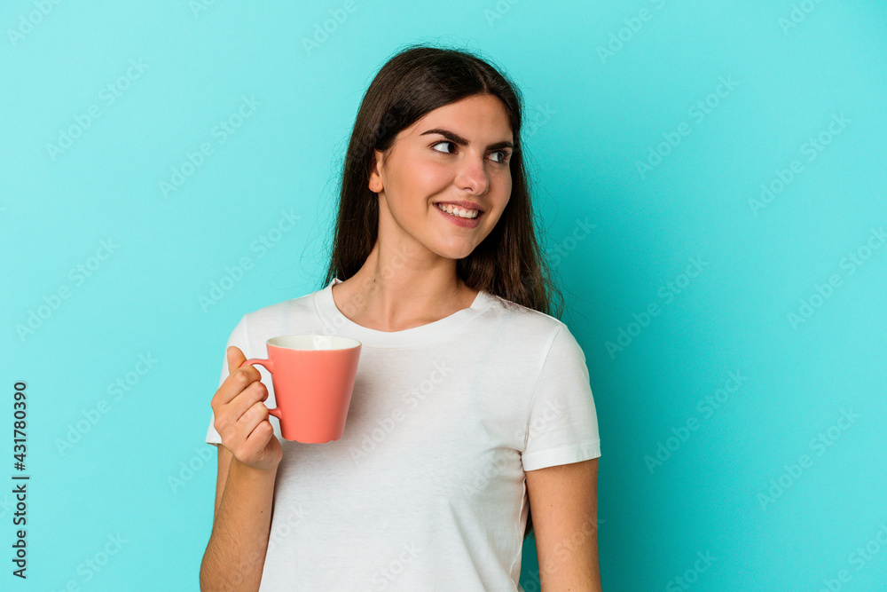 Young caucasian woman holding a mug isolated on blue background looks aside smiling, cheerful and pleasant. Young caucasian woman holding a mug isolated on blue background looks aside smiling, cheerful and pleasant.