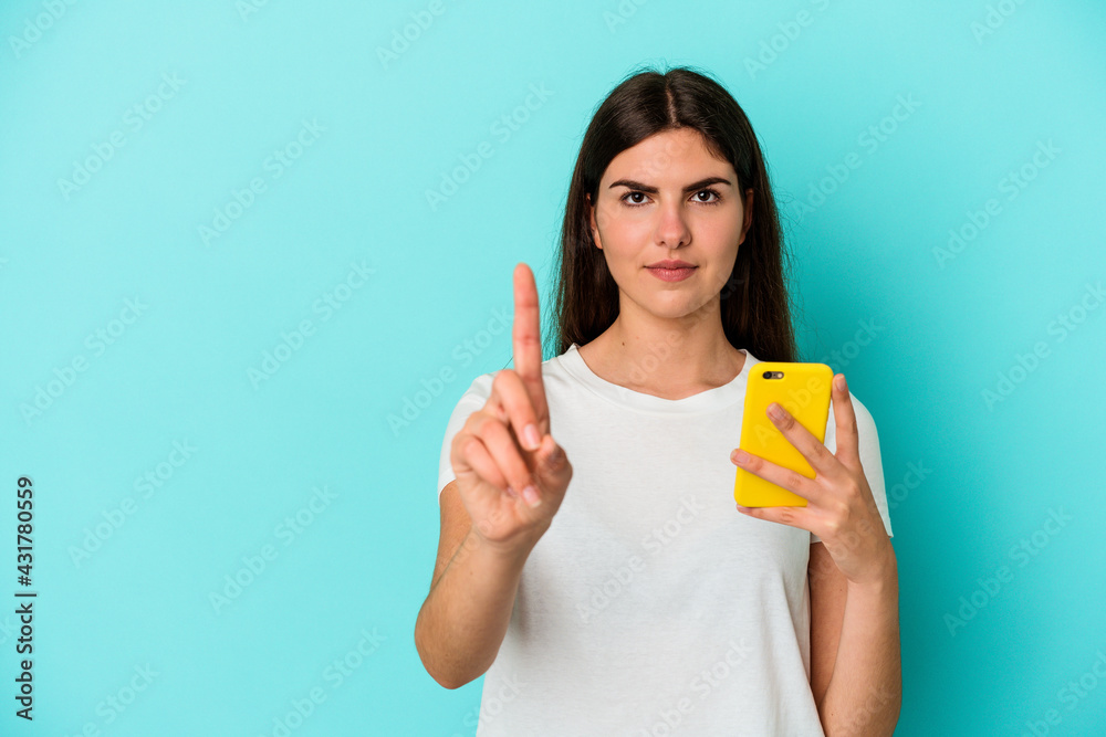 Young caucasian woman holding a mobile phone isolated on blue background showing number one with finger. Young caucasian woman holding a mobile phone isolated on blue background showing number one with finger.