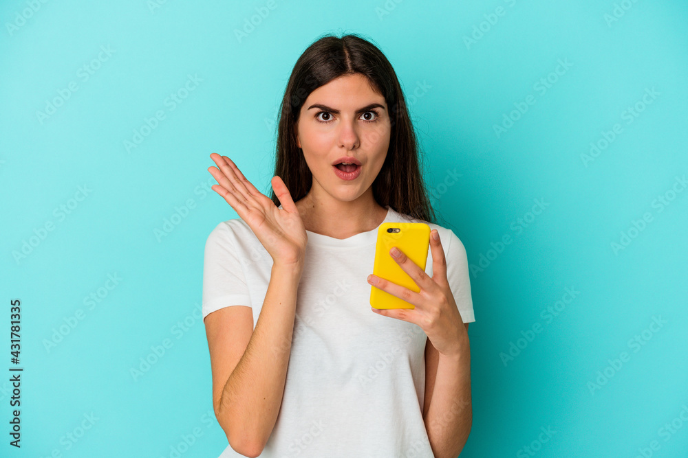 Young caucasian woman holding a mobile phone isolated on blue background surprised and shocked. Young caucasian woman holding a mobile phone isolated on blue background surprised and shocked.