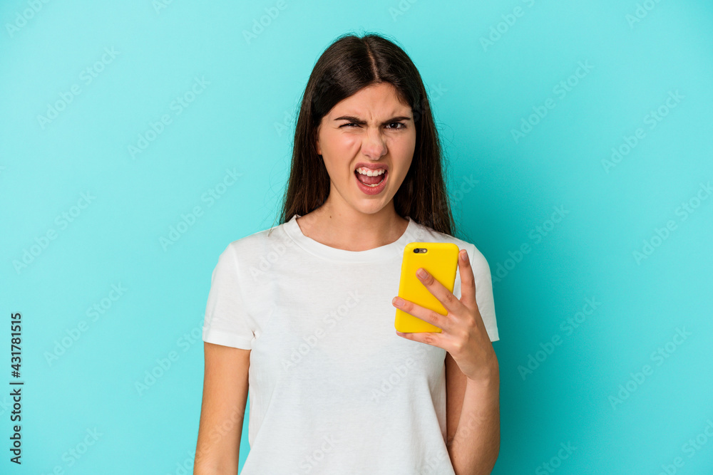 Young caucasian woman holding a mobile phone isolated on blue background screaming very angry and aggressive. Young caucasian woman holding a mobile phone isolated on blue background screaming very angry and aggressive.