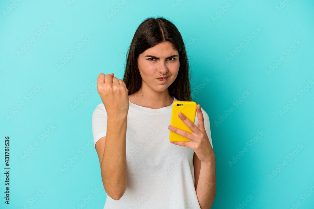 Young caucasian woman holding a mobile phone isolated on blue background showing fist to camera, aggressive facial expression.