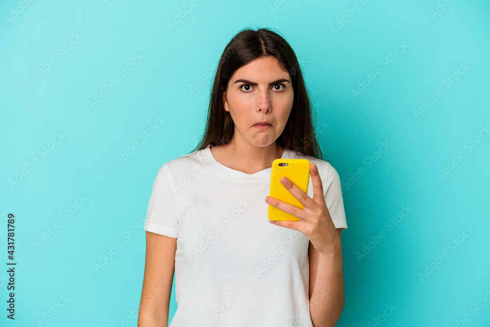 Young caucasian woman holding a mobile phone isolated on blue background shrugs shoulders and open eyes confused. Young caucasian woman holding a mobile phone isolated on blue background shrugs shoulders and open eyes confused.
