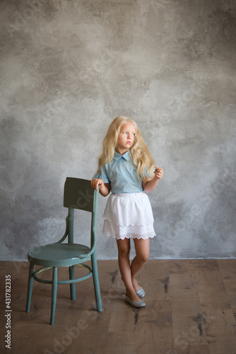 beautiful blonde girl stands near a chair against background of a gray wall. Photo demonstrating the character child - daydreaming, enthusiasm, vulnerability, touching, beauty, stubbornness, cunning