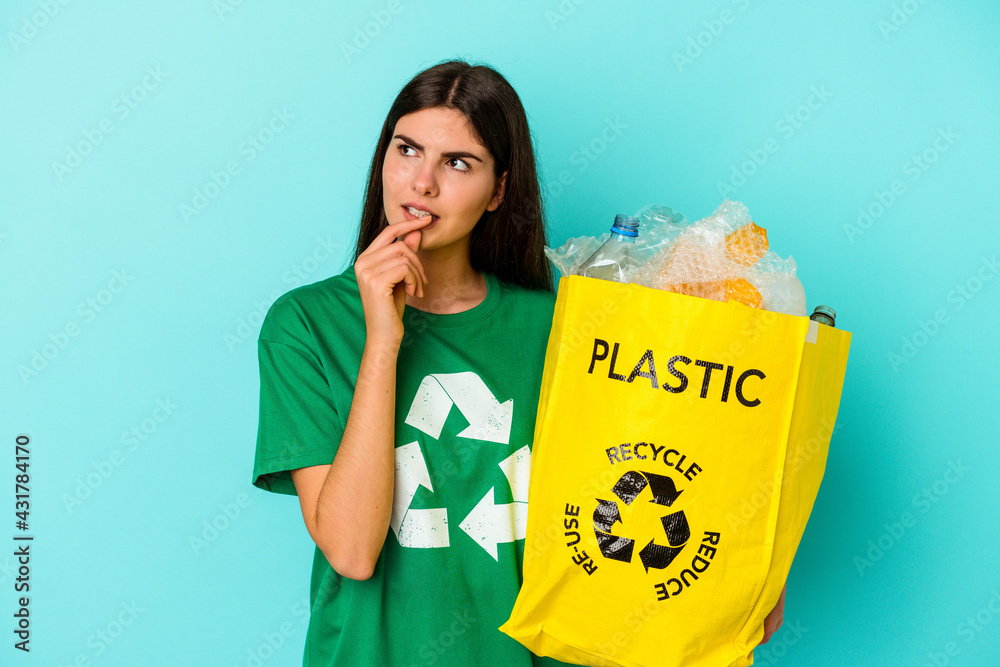 Young caucasian woman recycled plastic isolated on blue background relaxed thinking about something looking at a copy space. Young caucasian woman recycled plastic isolated on blue background relaxed thinking about something looking at a copy space.