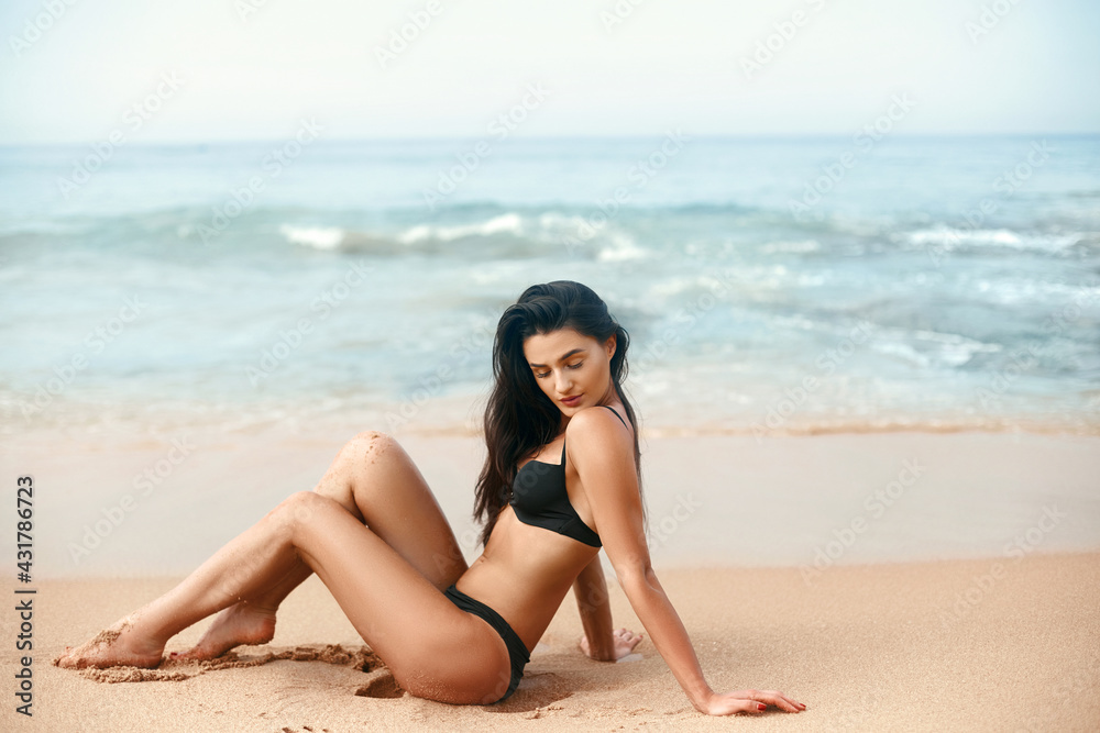 Fashion photo of beautiful happy slender woman sitting at sea. Beautiful luxury slim girl in a black bikini on the beach the ocean. Sexy tanned body.