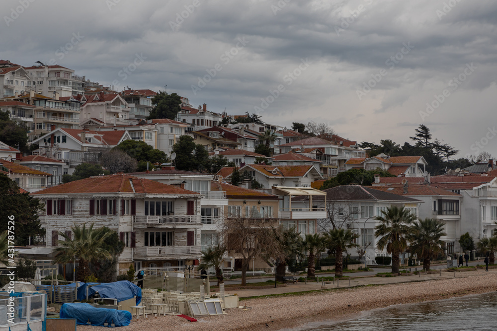 View from the sea on the island of Heybeliada in Istanbul. The slope shore is covered with many white summer cottages with brown tiled roofs. Beach with pebbles. Dramatic grey clouds in a stormy sky