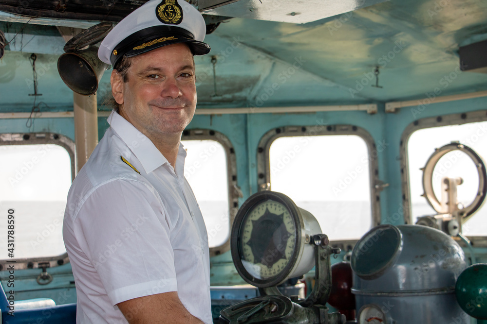 A captain standing in the wheelhouse of ship and looking to camera. - obrazy, fototapety, plakaty 