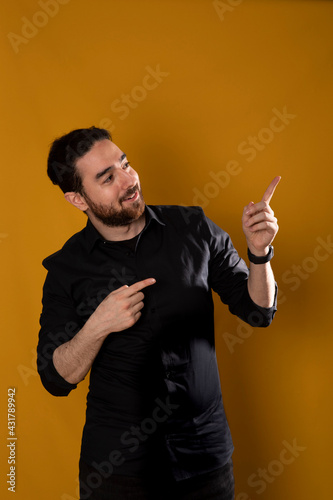 Südländischer junger Mann steht vom gelben Hintergrund und zeigt mit den Fingern oben rlinks. In der Richtung schauet er auch nach. Er ist schwarz bekleidet und hat schwarzen Bart.  Studio Foto.