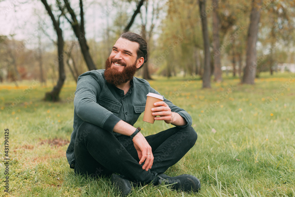 Portrait of smiling bearded man sitting in park on grass and drinking cup of coffee to go Portrait of smiling bearded man sitting in park on grass and drinking cup of coffee to go