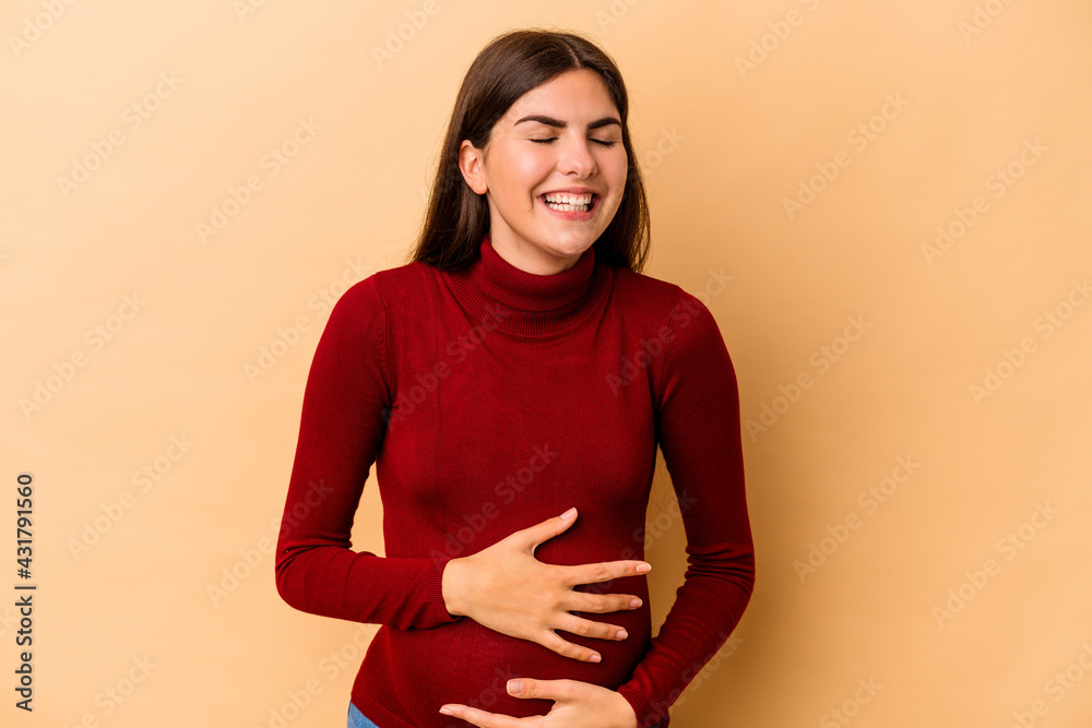 Obraz premium Young caucasian pregnant woman isolated on beige background laughs happily and has fun keeping hands on stomach.