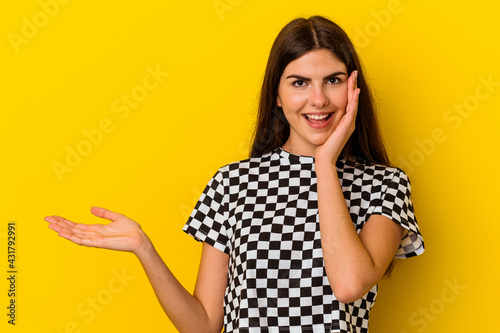 Young caucasian woman isolated on yellow background holds copy space on a palm, keep hand over cheek. Amazed and delighted. © Asier
