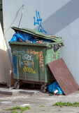 Duży zielony kosz na śmieci pomalowany farbą w sprayu. Tekst graffiti.