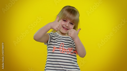 Little blonde teen kid child girl raises thumbs up agrees with something or gives positive reply recommends advertisement likes good posing isolated on yellow studio background. Young children emotion