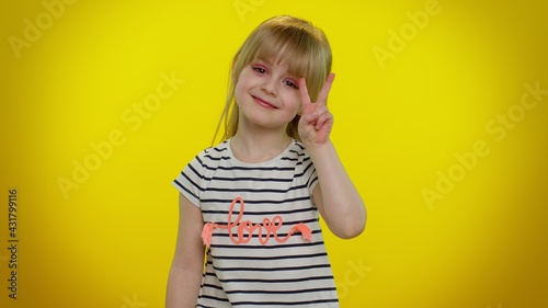 Funny blonde kid child 5-6 years old showing victory sign hoping for success and win, doing peace gesture, smiling with kind optimistic expression on yellow background. Teenager children girl emotions