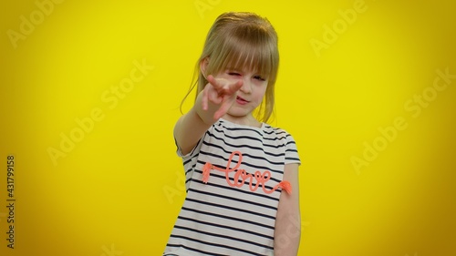 Little blonde teen child kid girl in t-shirt pointing at her eyes and camera, show I am watching you gesture, spying on someone. Young children posing on yellow studio background. Childhood lifestyle