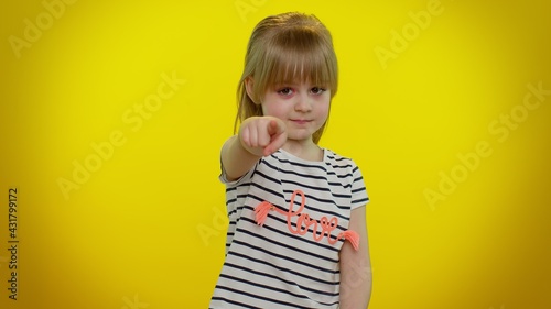 I choose you. Little blonde kid child girl pointing to camera and looking with playful happy expression, making choice, showing direction. Isolated on yellow studio background. Young children emotions