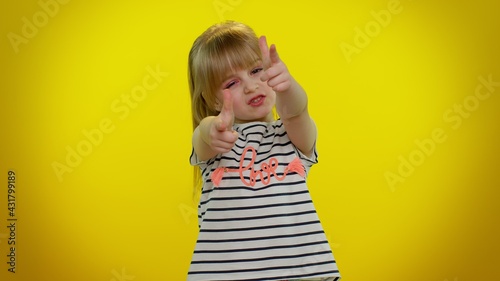 Little blonde teen kid child girl in t-shirt pointing around with finger gun gesture, looking confident, making choice, shooting killing with hand pistol right on target on yellow studio background
