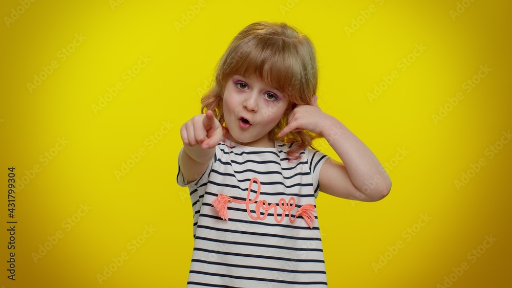 Little cute blonde teen child kid girl in white black striped t-shirt looking at camera doing phone gesture like says hey you call me back. Young children posing isolated on yellow studio background Little cute blonde teen child kid girl in white black striped t-shirt looking at camera doing phone gesture like says hey you call me back. Young children posing isolated on yellow studio background