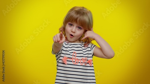 Little cute blonde teen child kid girl in white black striped t-shirt looking at camera doing phone gesture like says hey you call me back. Young children posing isolated on yellow studio background