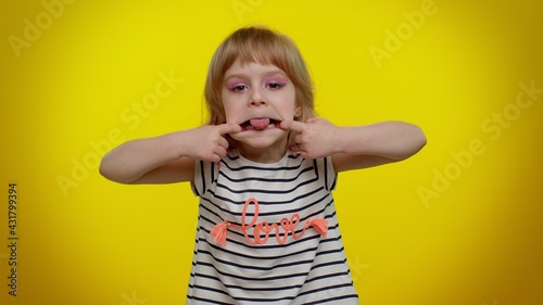 Portrait of cheerful funny blonde child girl 5-6 years old showing tongue making faces at camera, fooling around, joking, aping with silly face, teasing. Yellow studio background. Teen kid children