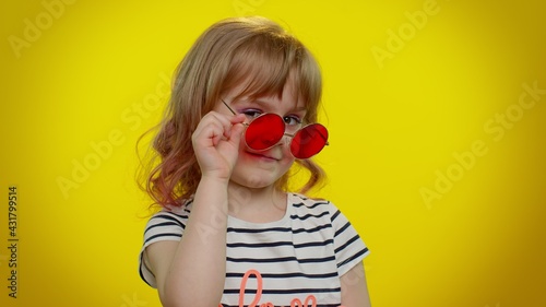 Portrait of seductive funny playful blonde kid child 5-6 years old wearing sunglasses, blinking eye, looking at camera with charming smile. Stylish teenager children girl on yellow studio background