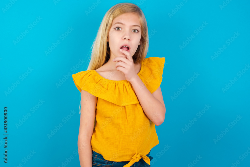 Nervous puzzled Caucasian kid girl wearing yellow T-shirt against blue wall opens mouth from surprise, reacts on sudden news. Nervous puzzled Caucasian kid girl wearing yellow T-shirt against blue wall opens mouth from surprise, reacts on sudden news.