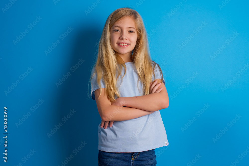 beautiful caucasian little girl wearing blue T-shirt over blue background being happy smiling and crossed arms looking confident at the camera. Positive and confident person.