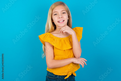 Optimistic Caucasian kid girl wearing yellow T-shirt against blue wall keeps hands partly crossed and hand under chin, looks at camera with pleasure. Happy emotions concept.