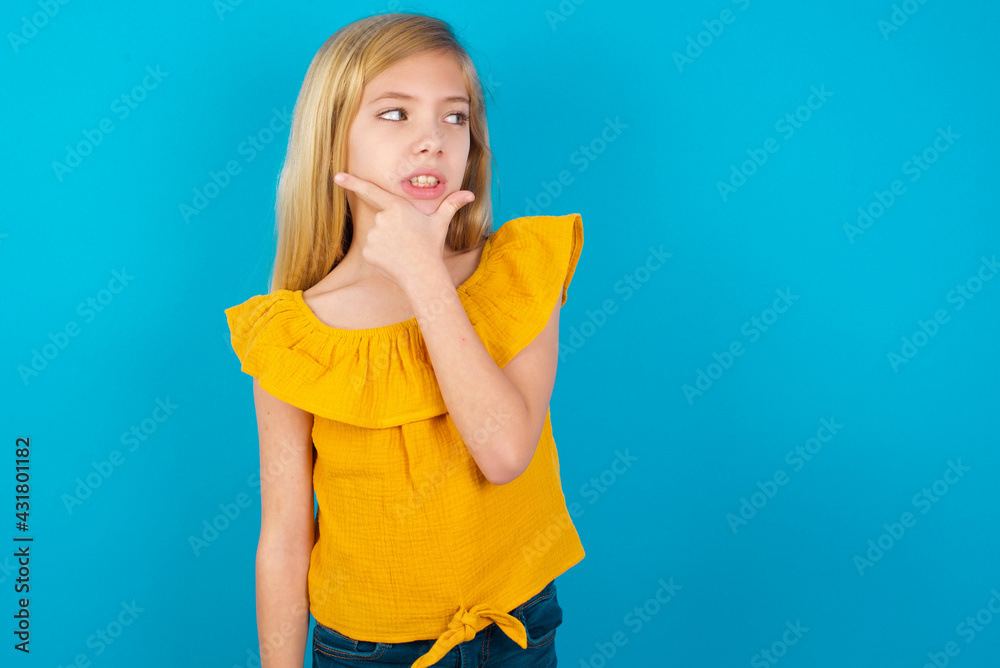 Caucasian kid girl wearing yellow T-shirt against blue wall Thinking worried about a question, concerned and nervous with hand on chin. Caucasian kid girl wearing yellow T-shirt against blue wall Thinking worried about a question, concerned and nervous with hand on chin.