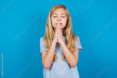 Indoor closeup of beautiful Caucasian little girl wearing white T-shirt over blue background practicing yoga and meditation, holding palms together in namaste, looking calm, relaxed and peaceful.