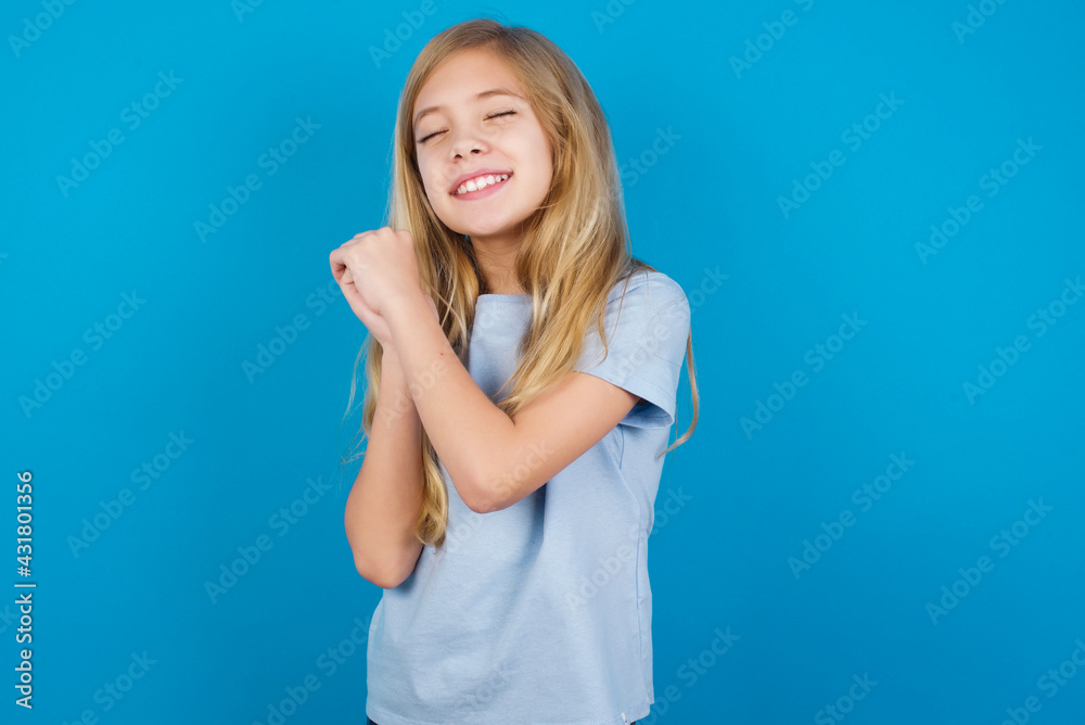 Dreamy beautiful Caucasian little girl wearing blue T-shirt over blue background with pleasant expression, closes eyes, keeps hands crossed near face, thinks about something pleasant Dreamy beautiful Caucasian little girl wearing blue T-shirt over blue background with pleasant expression, closes eyes, keeps hands crossed near face, thinks about something pleasant