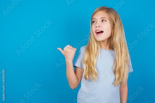 Stupefied beautiful Caucasian little girl wearing blue T-shirt over blu with surprised expression, opens eyes and mouth widely, points aside with thumb, shows something strange. Advertisement concept.