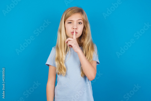 beautiful Caucasian little girl wearing blue T-shirt over blue background makes hush gesture, asks be quiet. Don't tell my secret or not speak too loud, please!
