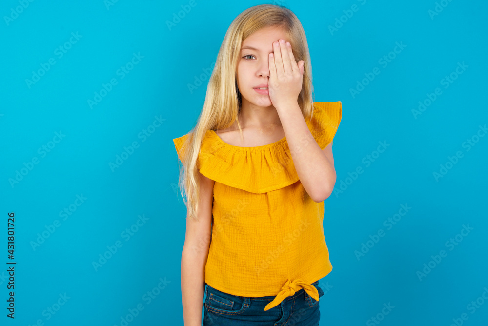 Caucasian kid girl wearing yellow T-shirt against blue wall covering one eye with her hand, confident smile on face and surprise emotion. Caucasian kid girl wearing yellow T-shirt against blue wall covering one eye with her hand, confident smile on face and surprise emotion.