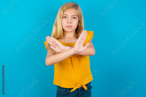 Caucasian kid girl wearing yellow T-shirt against blue wall has rejection expression crossing arms and palms doing negative sign, angry face.
