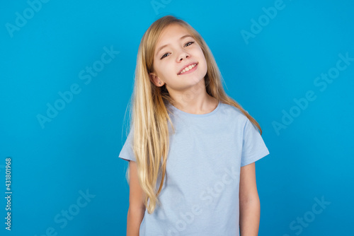 beautiful Caucasian little girl wearing blue T-shirt over blue background with broad smile, shows white teeth, feeling confident rejoices having day off. © Jihan