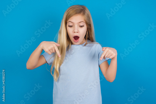 Billede på lærred Amazed beautiful Caucasian little girl wearing blue T-shirt over blue background points down with fore fingers, opens mouth being shocked