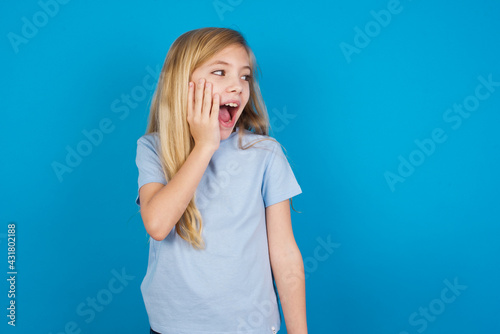 beautiful Caucasian little girl wearing blue T-shirt over blue background excited looking to the side hand on face. Advertisement and amazement concept.