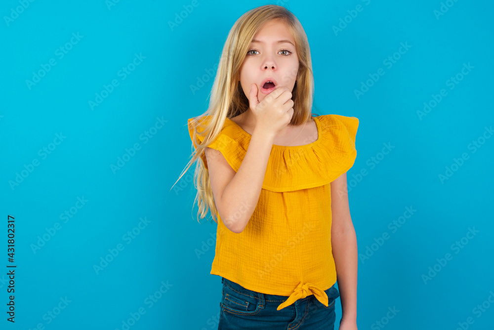 Caucasian kid girl wearing yellow T-shirt against blue wall Looking fascinated with disbelief, surprise and amazed expression with hands on chin Caucasian kid girl wearing yellow T-shirt against blue wall Looking fascinated with disbelief, surprise and amazed expression with hands on chin