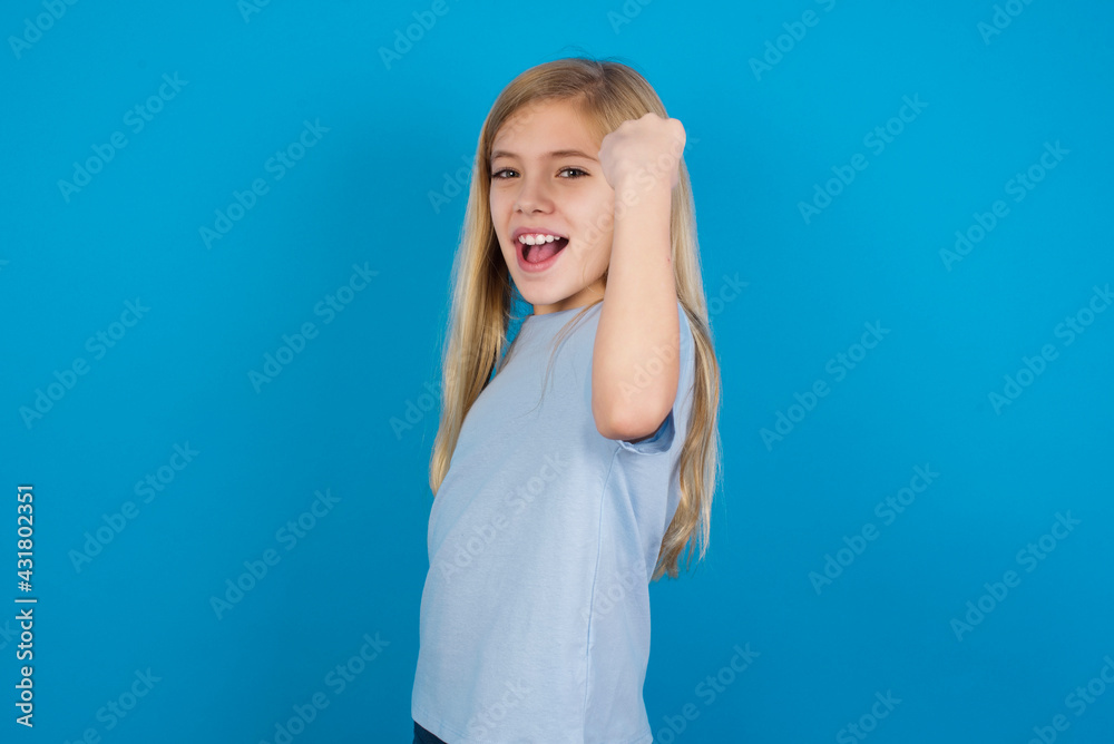 Fototapeta premium Overjoyed beautiful Caucasian little girl wearing blue T-shirt over blue background glad to receive good news, clenching fist and making winning gesture.