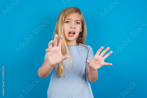 Fotografija Dissatisfied beautiful Caucasian little girl wearing white T-shirt over blue background frowns face, has disgusting expression, shows tongue, expresses non compliance, irritated with somebody