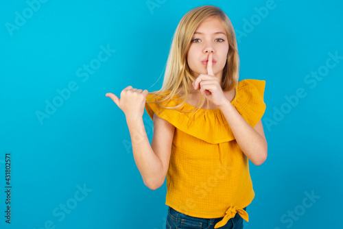 Caucasian kid girl wearing yellow T-shirt against blue wall asking to be quiet with finger on lips pointing with hand to the side. Silence and secret concept.