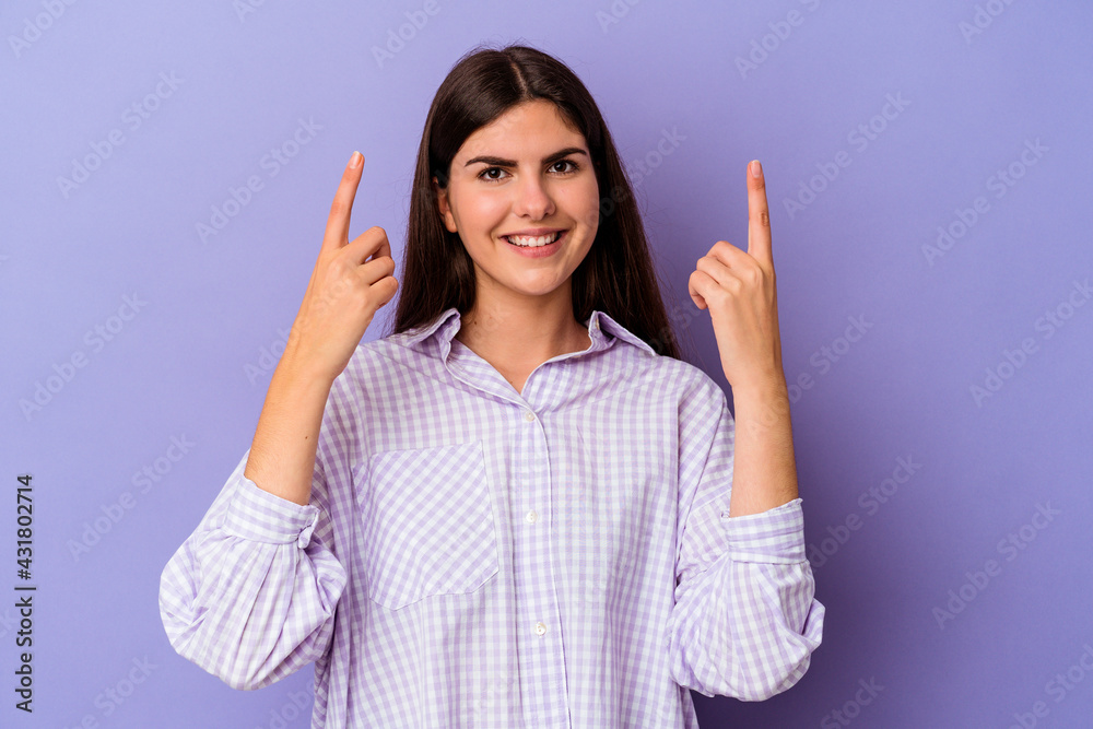 Young caucasian woman isolated on purple background indicates with both fore fingers up showing a blank space. Young caucasian woman isolated on purple background indicates with both fore fingers up showing a blank space.