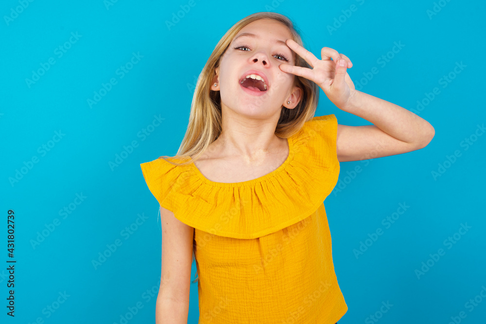 Caucasian kid girl wearing yellow T-shirt against blue wall Doing peace symbol with fingers over face, smiling cheerful showing victory Caucasian kid girl wearing yellow T-shirt against blue wall Doing peace symbol with fingers over face, smiling cheerful showing victory
